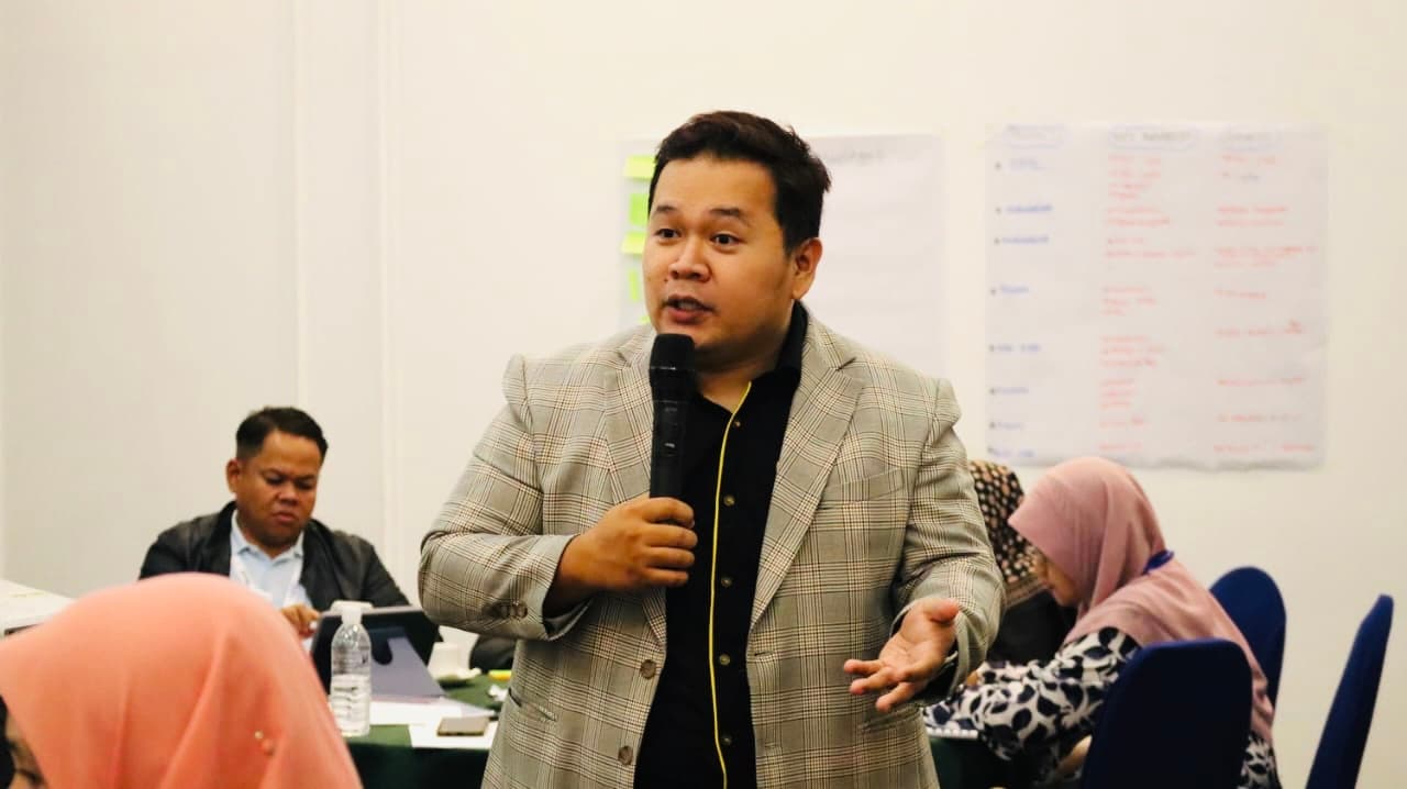 A man in a grey plaid blazer speaks into a microphone during a professional workshop.