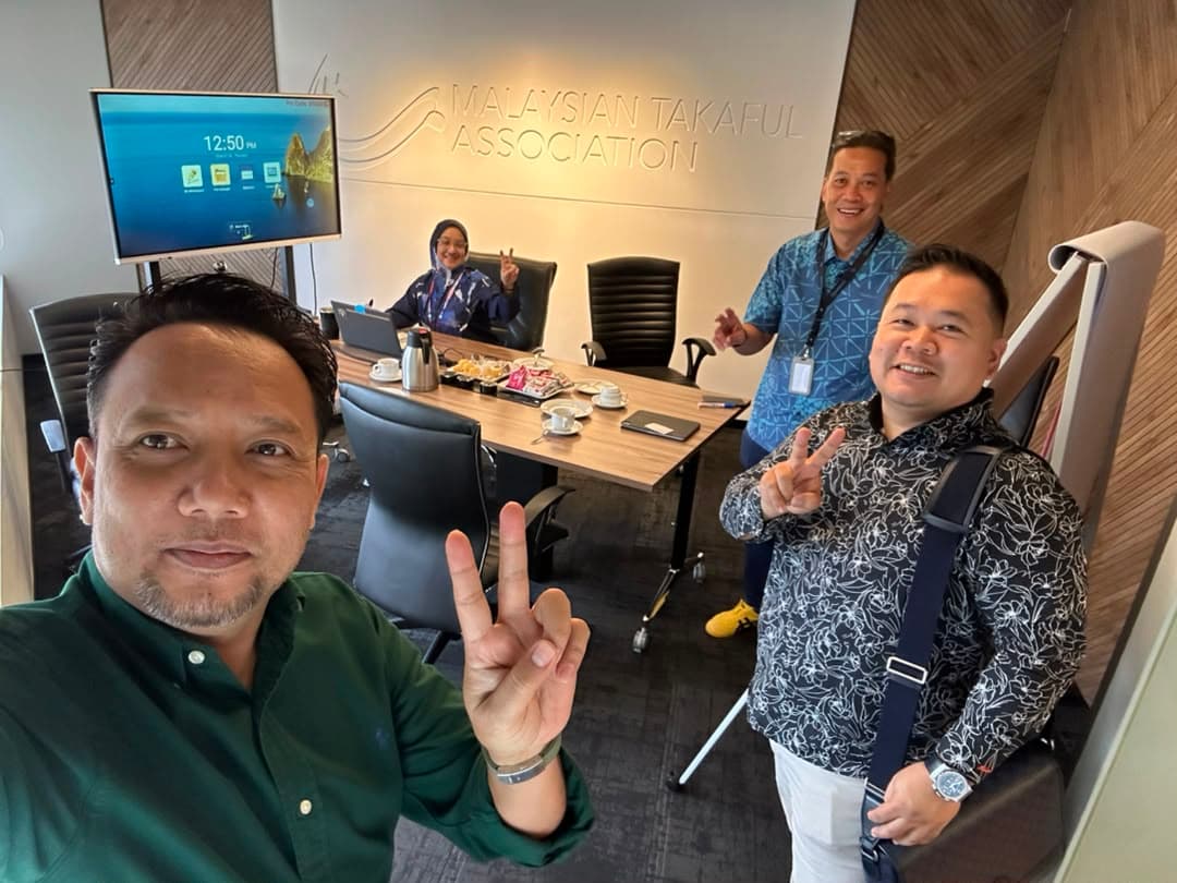 Four smiling colleagues pose for a selfie at the Malaysian Takaful Association office, making peace signs.